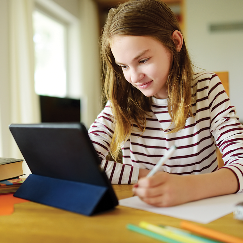 A focused child, engaged in a creative writing project, writes on paper while referring to a tablet for inspiration and research.