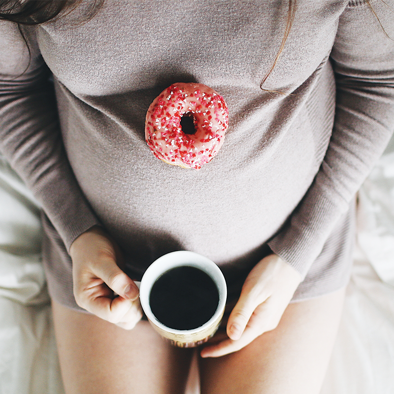 A radiant pregnant woman, cradling her growing belly, indulges in a sweet treat, a donut, while sipping on a warm cup of coffee.