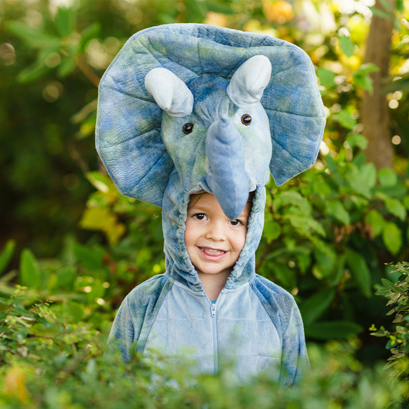 A playful child, dressed in a dinosaur costume standing behind a bush.