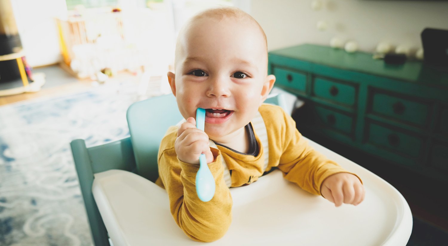 A curious baby, sitting in a high chair, explores a spoon, playfully holding it in their mouth.