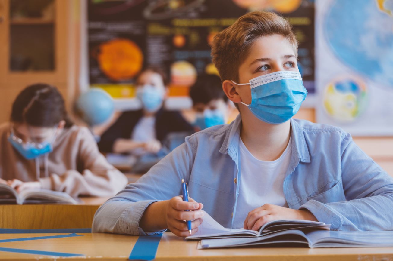 A young student, wearing a protective face mask, sits at their desk, focused on their studies, demonstrating the importance of safety measures in educational settings.