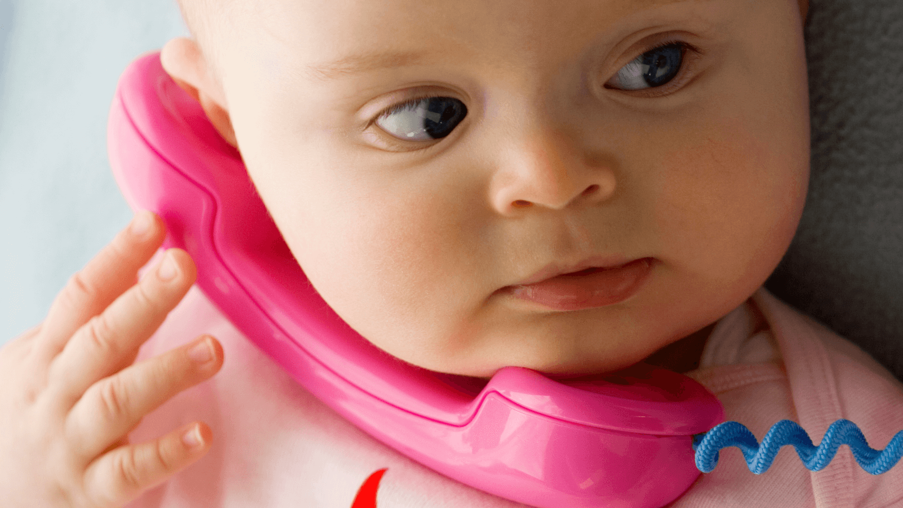 A baby holding a pink toy telephone to their ear, representing early speech development milestones.