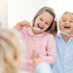 Two happy children, a boy and a girl, laughing while brushing their teeth in a bathroom mirror.