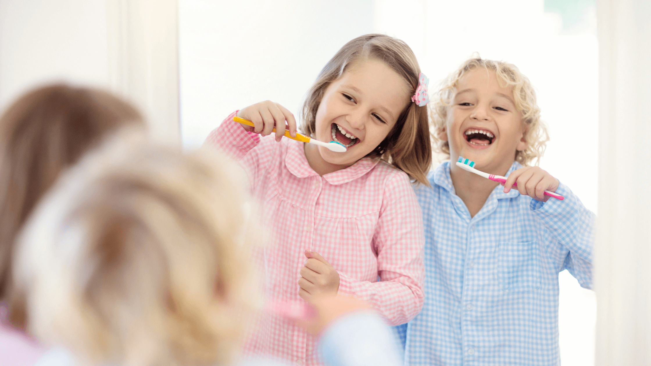 Two happy children, a boy and a girl, laughing while brushing their teeth in a bathroom mirror.