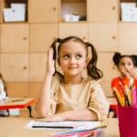 A young girl with pigtails raises her hand in a brightly lit classroom with other students.