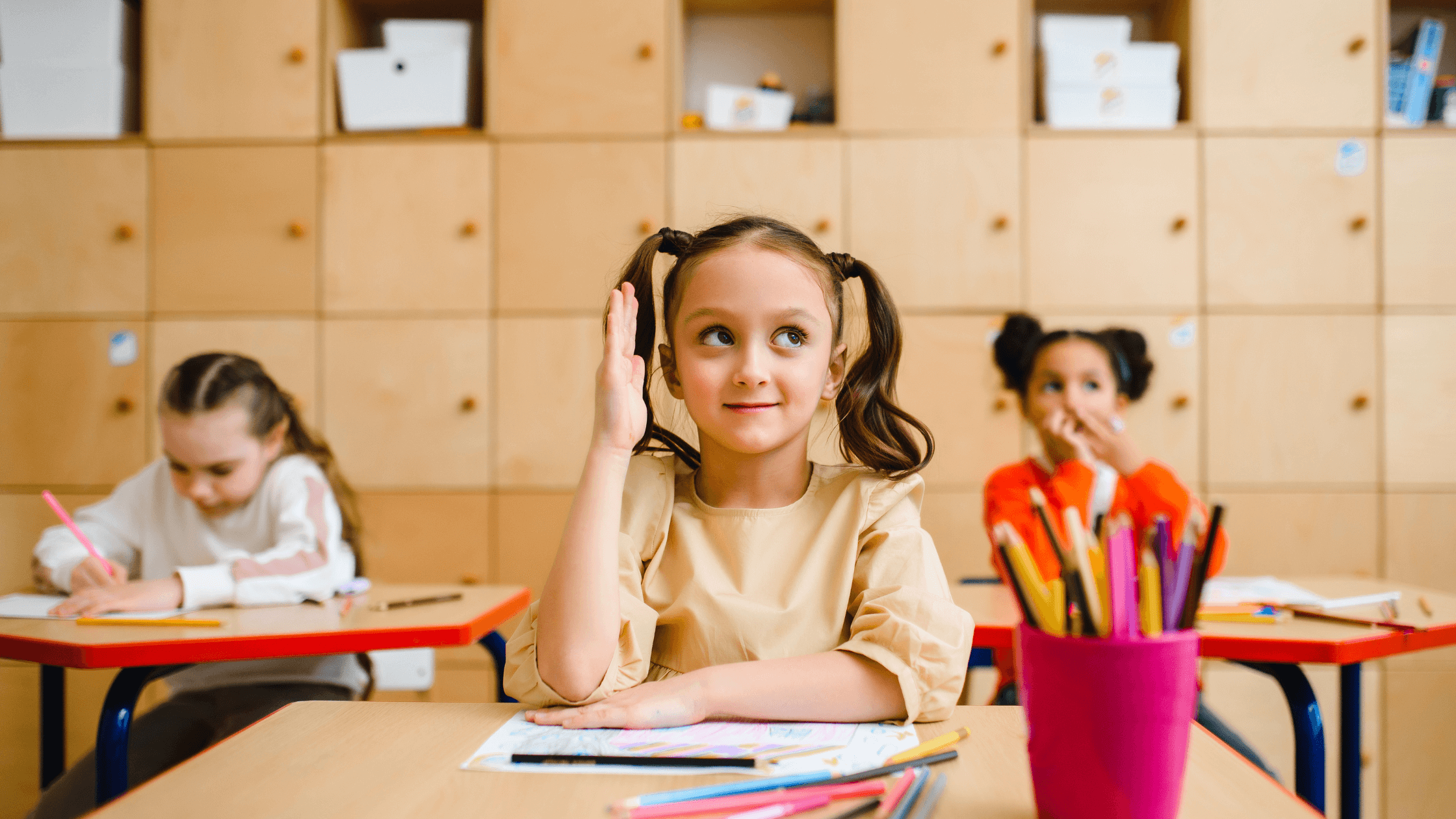 A young girl with pigtails raises her hand in a brightly lit classroom with other students.