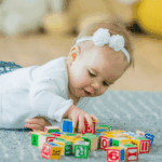 An adorable baby with a white headband is on their tummy on a gray rug, reaching for colorful alphabet blocks.