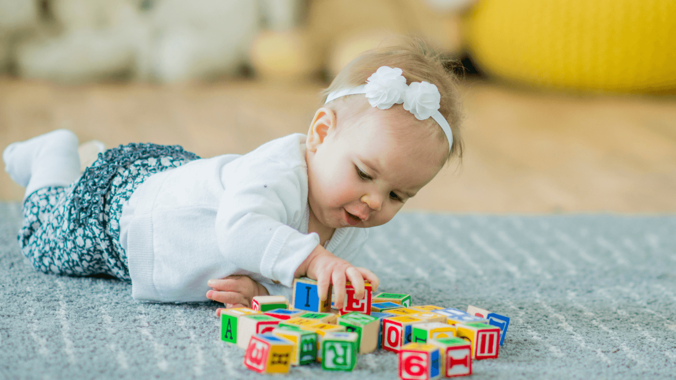 An adorable baby with a white headband is on their tummy on a gray rug, reaching for colorful alphabet blocks.