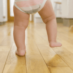 A baby's chubby legs and feet are shown from behind as they take a step on a wooden floor, wearing a diaper.