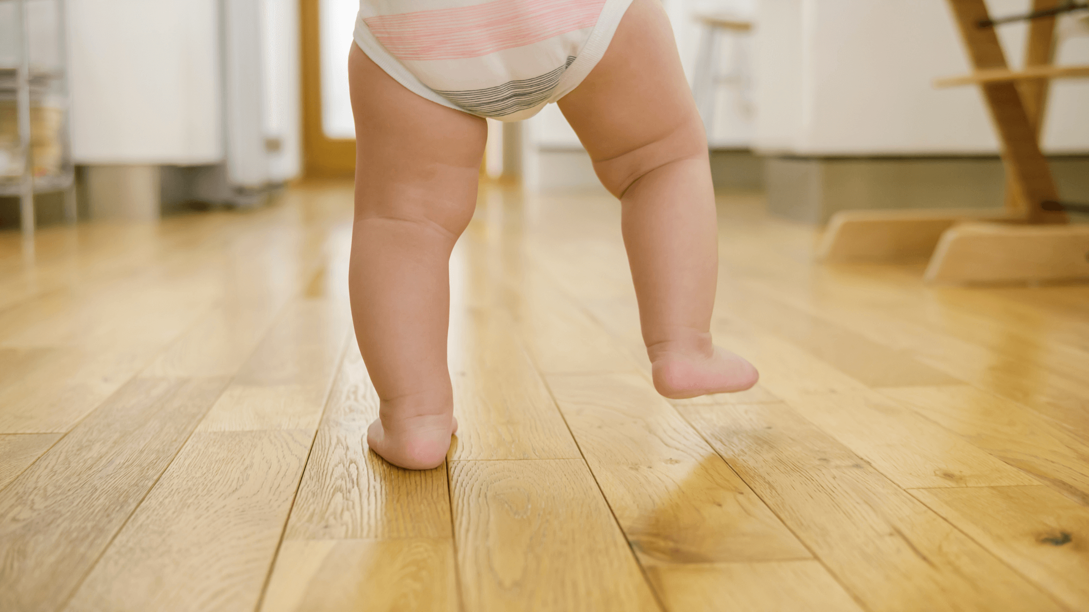 A baby's chubby legs and feet are shown from behind as they take a step on a wooden floor, wearing a diaper.